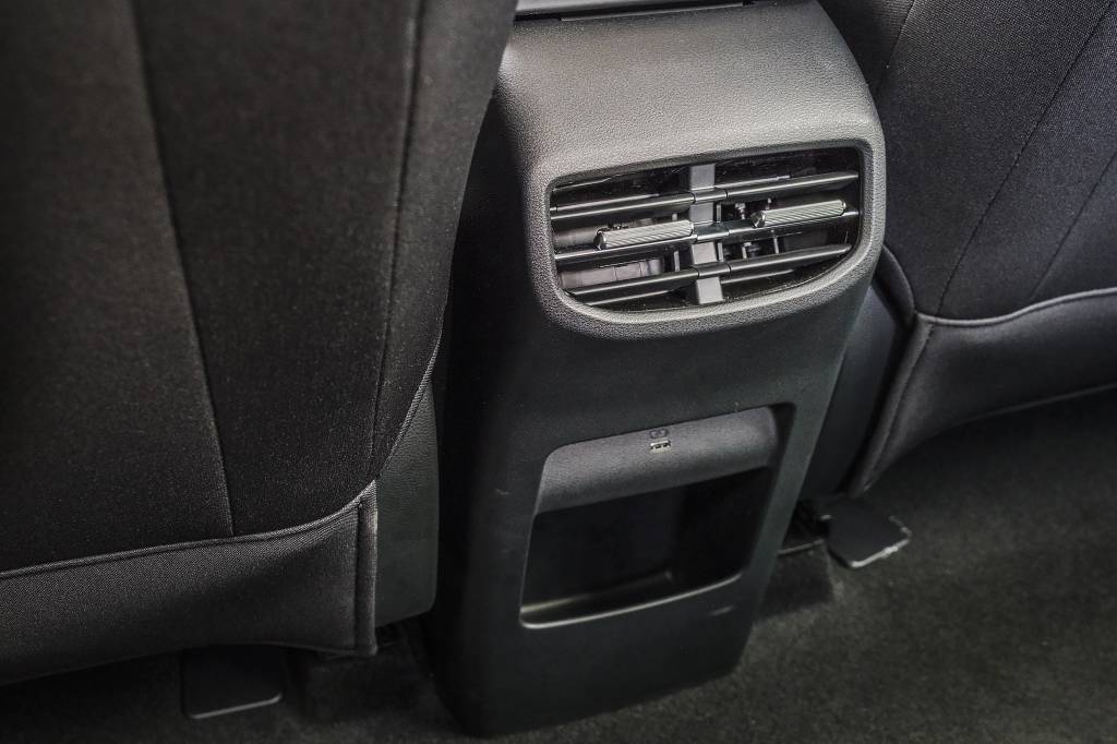 mg4-urban-quatro-rodas (47) Saída de ar condicionado traseira em um carro, com uma porta USB e um pequeno porta-objetos abaixo, entre dois bancos pretos