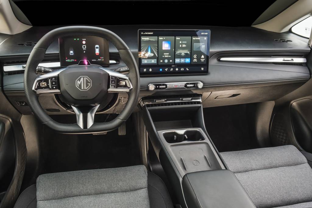 mg4-urban-quatro-rodas (40) Interior moderno de um carro, com volante multifuncional MG, painel digital e tela multimídia central exibindo aplicativos. Bancos cinza claro e console central com porta-copos.