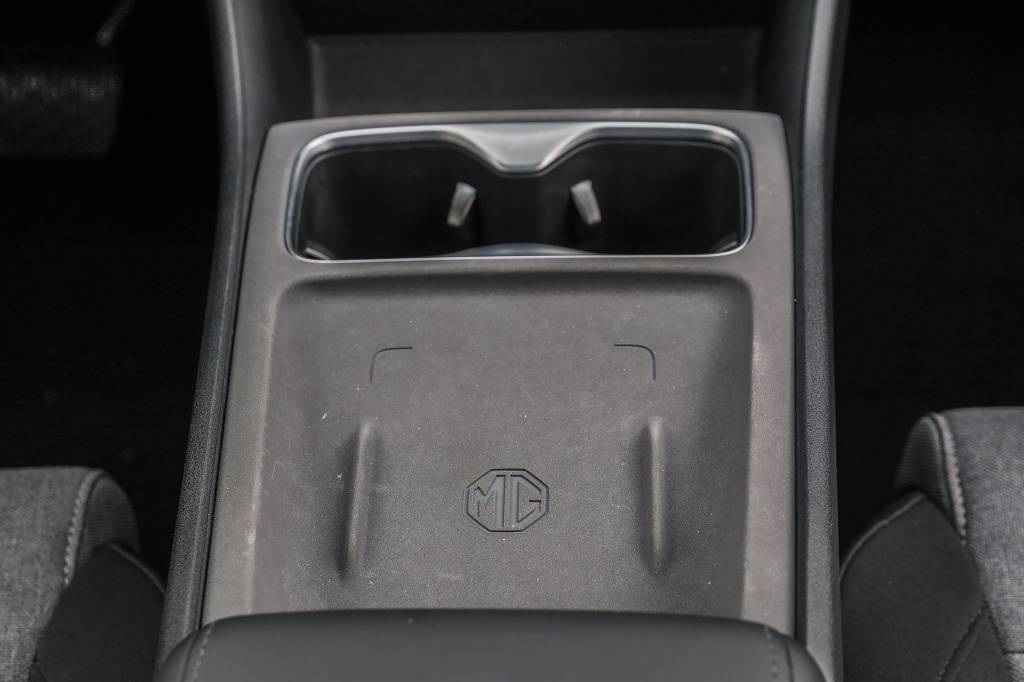 mg4-urban-quatro-rodas (21) Console central de carro cinza escuro com dois porta-copos cromados e um compartimento de carregamento sem fio com o logotipo MG em relevo