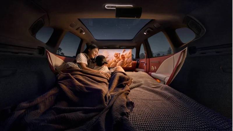Mãe e filha assistem a um filme em um carro adaptado para acampamento, com o banco traseiro transformado em cama e um projetor exibindo a imagem na parte de trás. O teto solar mostra o céu noturno estrelado, criando uma atmosfera aconchegante e de aventura