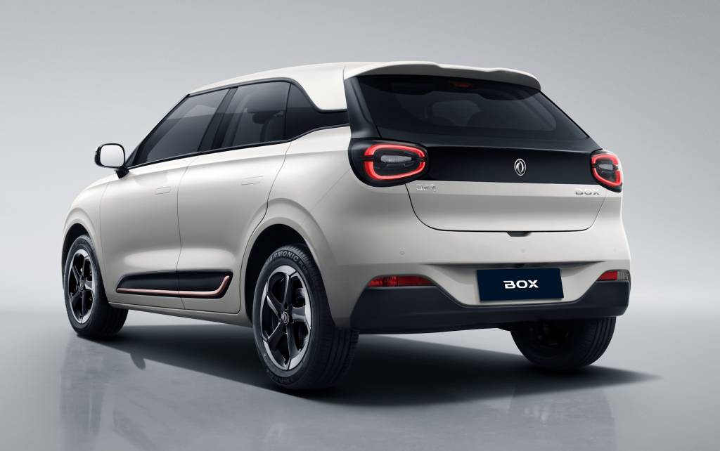Carro elétrico branco, modelo SUV compacto, visto de traseira e lateral direita, com detalhes em preto na tampa do porta-malas e nas rodas. As lanternas traseiras são em formato de C invertido e a placa exibe BOX em branco sobre fundo azul escuro. O veículo está em um estúdio com fundo cinza claro e reflexo no chão