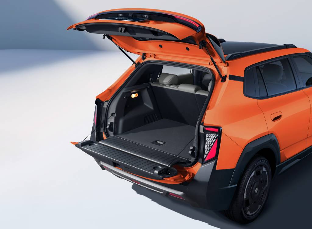 Traseira de um SUV laranja com a tampa do porta-malas aberta, revelando um amplo espaço interno e uma prateleira retrátil. As lanternas traseiras têm design moderno e o para-choque é preto com detalhes prateados. O veículo está em um estúdio com fundo claro