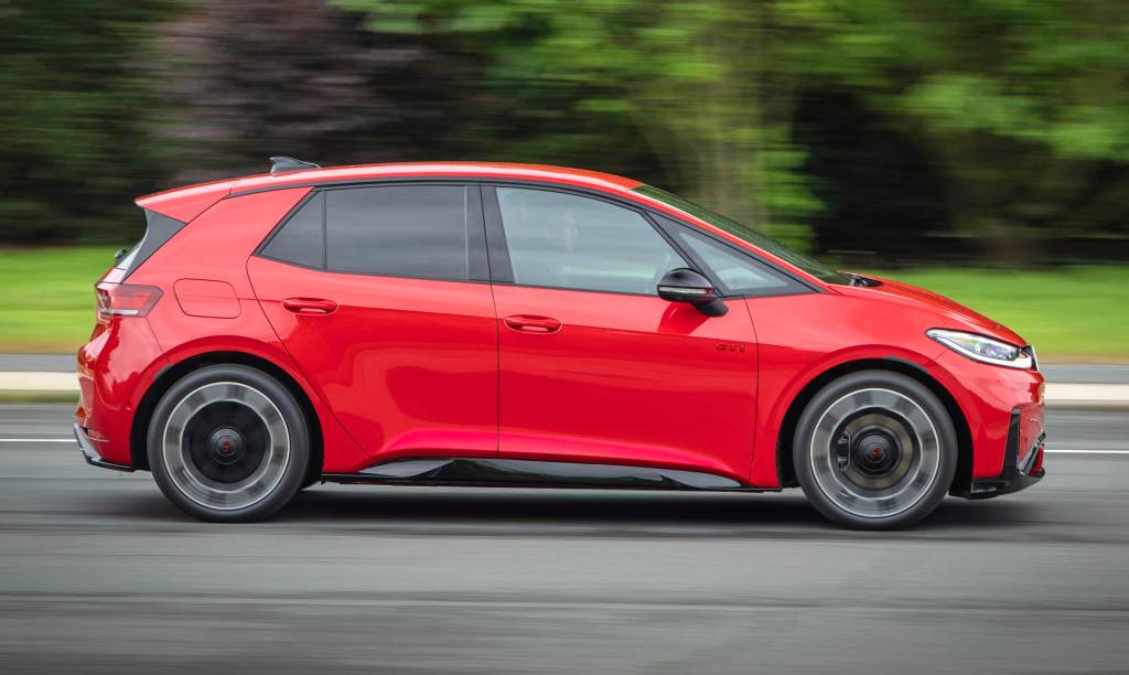 Carro compacto vermelho em movimento, visto de lado, com rodas girando rapidamente e fundo borrado de árvores verdes