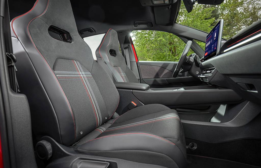 Interior de carro com bancos esportivos cinza-escuros e detalhes em vermelho, costuras vermelhas e padrão xadrez. Volante e painel pretos, tela multimídia vertical no centro. Vista externa de árvores verdes pela janela.