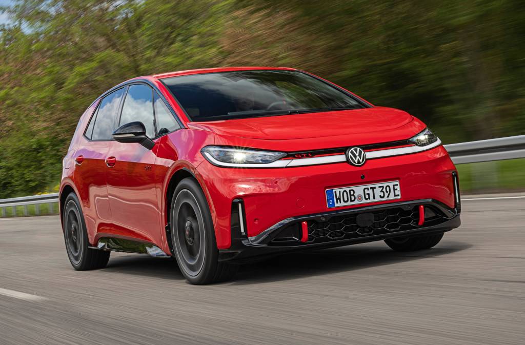 Carro vermelho Volkswagen ID.3 GTI em movimento, visto de frente e levemente de lado, em uma estrada com vegetação borrada ao fundo. O veículo tem faróis acesos, grade frontal preta com detalhes vermelhos e placa alemã WOB GT 39E
