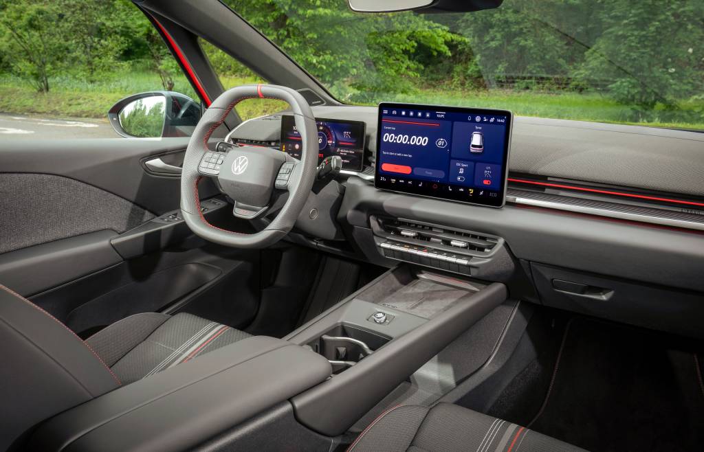 Interior de um carro moderno, com volante cinza e detalhes vermelhos, painel digital e uma tela multimídia grande exibindo um cronômetro e informações do veículo. O banco do motorista é cinza escuro com listras vermelhas e brancas. A paisagem verde de árvores é visível através do para-brisa e da janela lateral