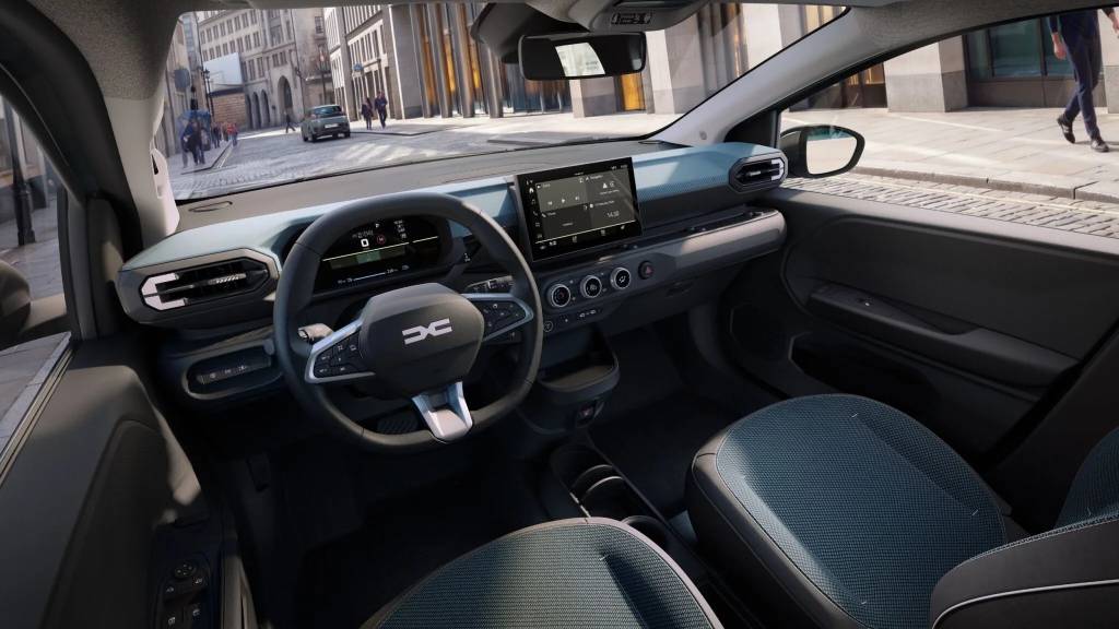 2027-Dacia-Spring-Interior-1-2048×1152 Interior de um carro moderno com volante multifuncional exibindo o logotipo Dacia, painel digital e tela multimídia central. O banco do passageiro tem estofamento azul e preto. Pelo para-brisa, vê-se uma rua urbana com prédios e pessoas caminhando.