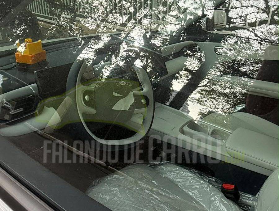 Omoda QQ em testes no Brasil Visão através da janela de um carro novo, com o interior branco e assentos cobertos por plástico. O volante é visível, e a paisagem externa reflete no vidro, mostrando árvores e folhagens. O texto FALANDO DE CARRO está sobreposto na imagem