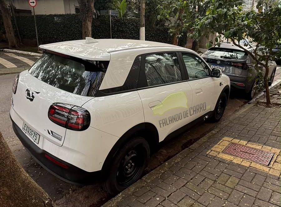 Omoda QQ em testes no Brasil Carro branco com adesivo FALANDO DE CARRO na lateral, estacionado em rua arborizada com calçada de blocos de concreto e outro veículo cinza ao fundo